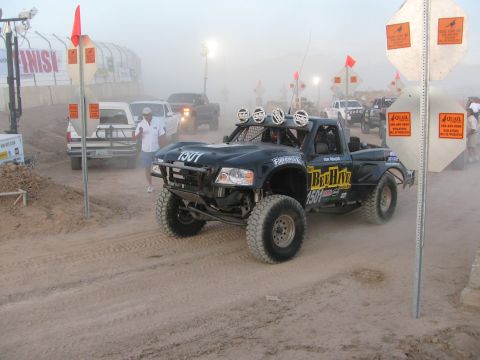 1990 Ford Bronco - black number 1501 Ford Ranger off-road race truck with roof lights moves through dust past flagged barriers at dusk.