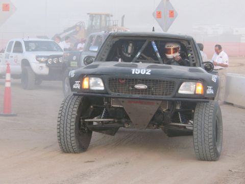 1990 Ford Bronco - black number 1502 off-road race truck rolls head-on through dust, its helmeted driver visible behind the roll cage.