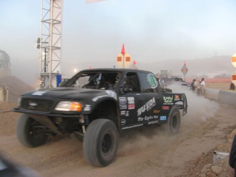1990 Ford Bronco - black Ford off-road race truck with Nu-Era sponsor decals throws up dust at a dusky staging area beside concrete barriers.
