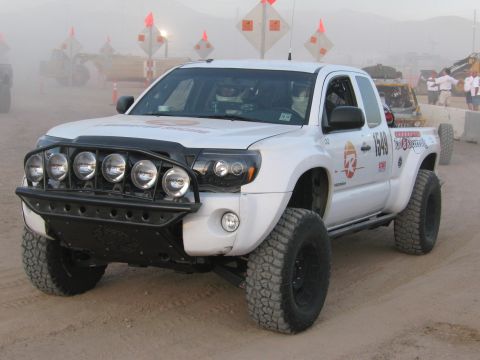 1990 Ford Bronco - white Toyota Tacoma prerunner numbered 1649 with a row of front-bumper light pods sits on a dusty desert course.