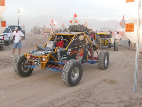1990 Ford Bronco - open tube-frame off-road buggy with 'Safari Cup' graphics and a spare tire idles at a dusty staging area amid spectators.