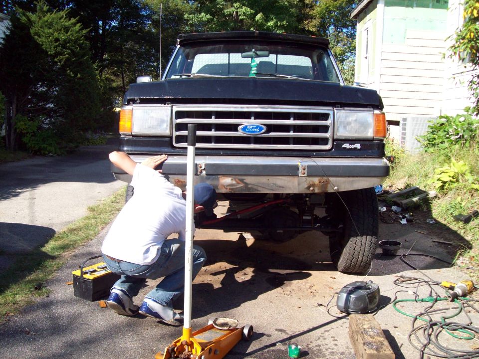 Person working under the front bumper of a black Ford F-150 pickup truck. Tools and a battery are on the ground
