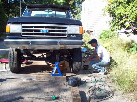 1989 Ford F-150 - A person works on the front end of a lifted Ford pickup truck that is supported by jack stands.