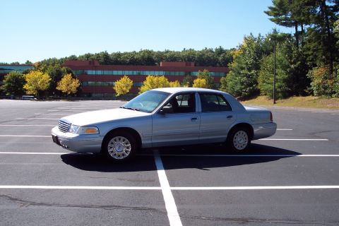 2001 Ford Crown Victoria - A silver 2001 Ford Crown Victoria sedan is parked in a lot with a building and trees in the background.