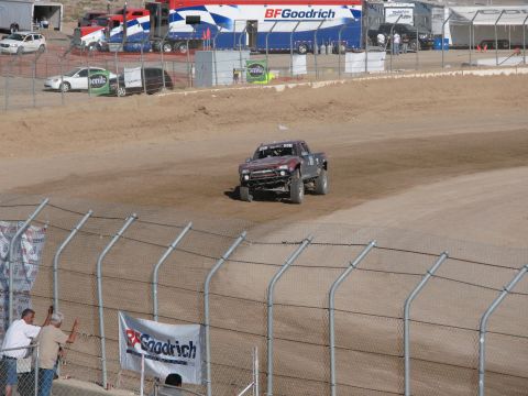 1990 Ford Bronco - dark off-road race truck rounds a banked dirt oval track behind a chain-link fence with BFGoodrich banners overhead.