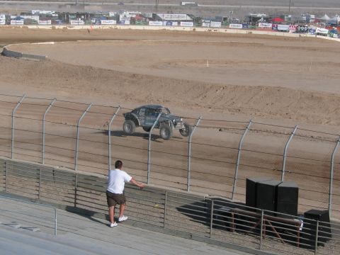 1990 Ford Bronco - spectator leans on a rail watching a tube-frame off-road racer on a dirt oval track from the grandstand.