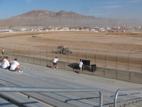 1990 Ford Bronco - Spectators in a grandstand watch a distant tube-frame off-road racer on a desert dirt oval beneath a barren mountain.