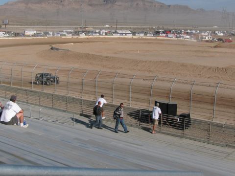 1990 Ford Bronco - Fans walk along a grandstand rail while a tube-frame off-road racer runs the far side of a desert dirt oval.