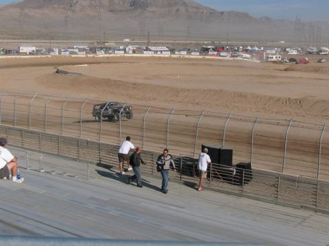1990 Ford Bronco - dark off-road race truck runs a desert dirt oval as a few spectators stroll the grandstand in the foreground.
