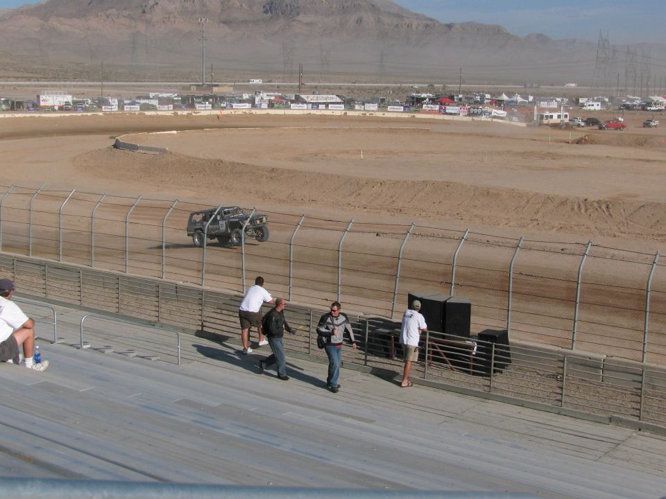 Dark off-road race truck on dirt track with spectators walking grandstand