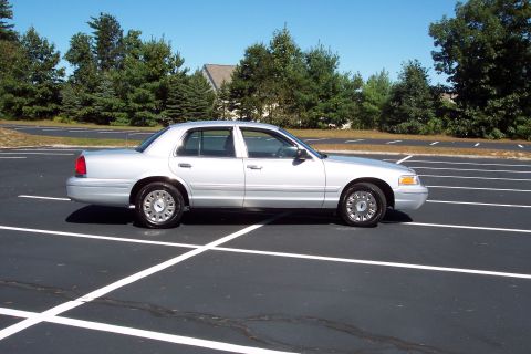 2001 Ford Crown Victoria - Side profile of a silver 2001 Ford Crown Victoria sedan parked in an empty lot under a clear blue sky.