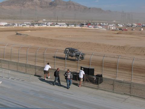 1990 Ford Bronco - dark off-road race truck runs the far bank of a desert dirt oval while spectators gather at the grandstand rail.