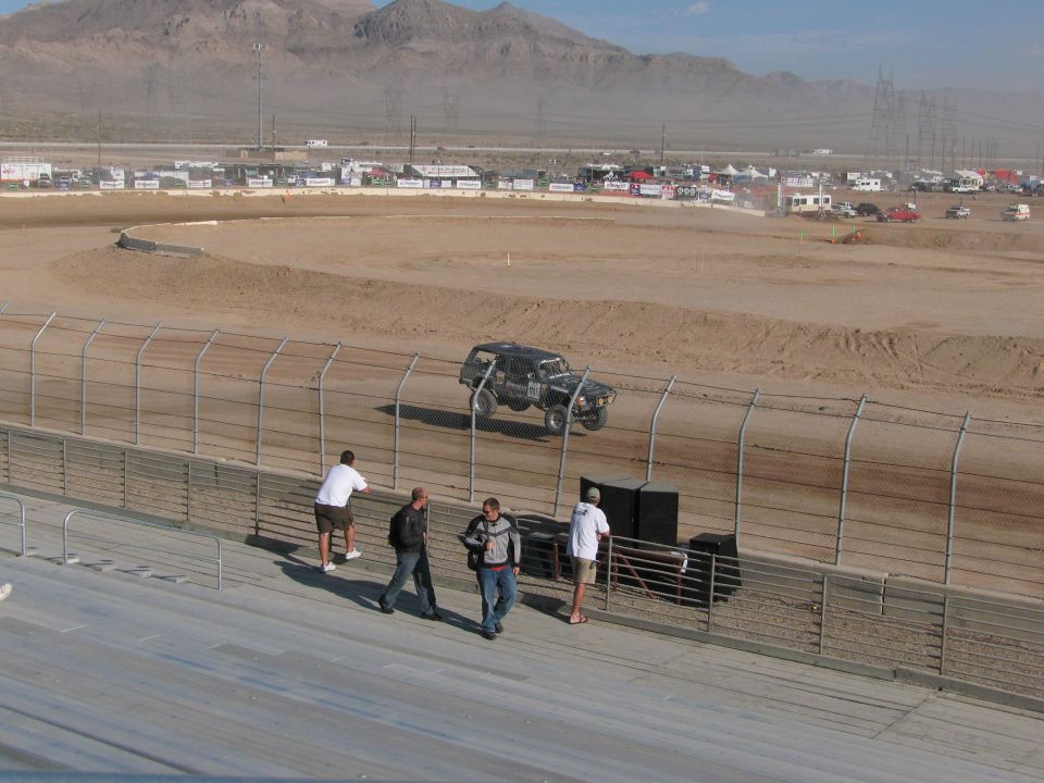 Dark off-road race truck on far dirt oval with fans at grandstand rail