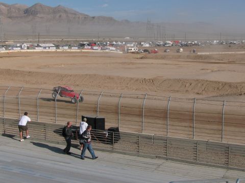 1990 Ford Bronco - red off-road racer runs a banked desert dirt oval while fans walk along the grandstand in the foreground.