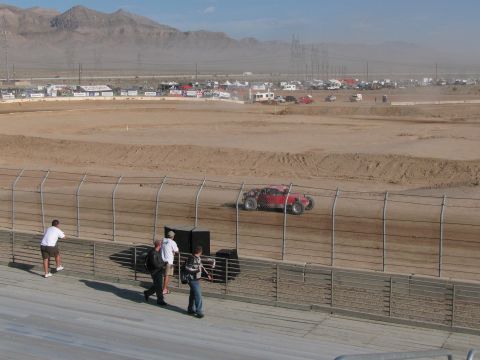 1990 Ford Bronco - red off-road racer rounds a desert dirt oval seen from a grandstand where a few spectators stand at the rail.