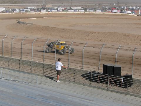 1990 Ford Bronco - yellow tube-frame off-road racer runs a desert dirt oval behind a chain-link fence as a spectator watches.