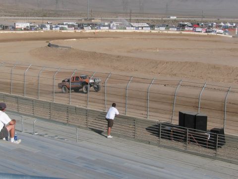 1990 Ford Bronco - orange off-road race truck runs a desert dirt oval as a few spectators watch from the grandstand rail.