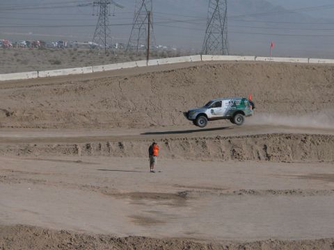 1990 Ford Bronco - silver-and-green off-road racer crests a sculpted dirt berm trailing dust as an orange-vested flagger watches.