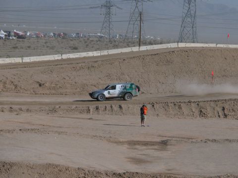 1990 Ford Bronco - silver-and-green off-road racer runs a sandy dirt course past an orange-vested flagger under power lines.