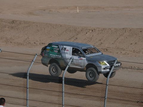 1990 Ford Bronco - silver number 878 off-road race SUV with charity decals catches air on a dirt course behind a chain-link fence.