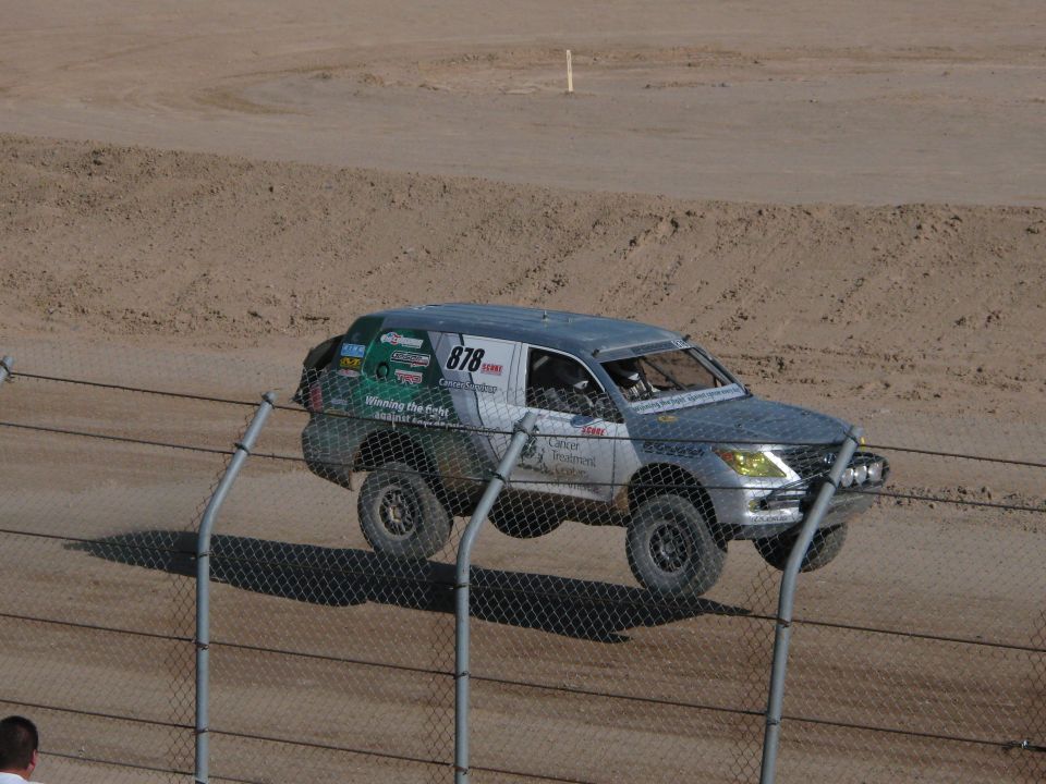 Silver number 878 off-road race SUV jumping behind chain-link fence