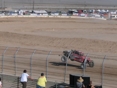 1990 Ford Bronco - red tube-frame off-road race truck catches air over a dirt course as spectators watch from behind a chain-link fence.