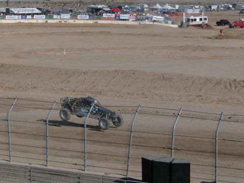 1990 Ford Bronco - dark-colored off-road race buggy rounds the banked dirt track past a chain-link fence at an outdoor racing venue.