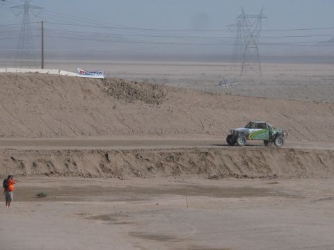 1990 Ford Bronco - green and white off-road race truck drives a dirt course past a BFGoodrich banner with transmission towers in the desert behind.
