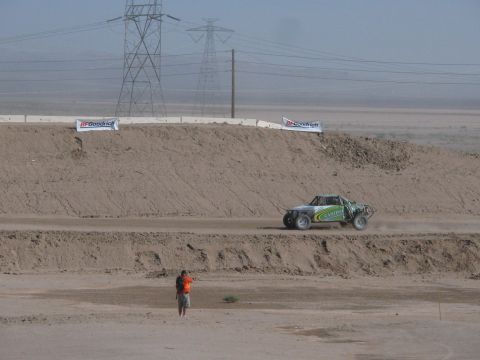 1990 Ford Bronco - green off-road race truck navigates a dirt berm as a marshal in orange points, with power transmission towers beyond.