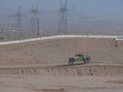 1990 Ford Bronco - green off-road race truck rounds a high dirt-walled corner beneath a row of electrical transmission towers in the desert.