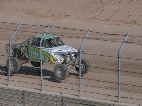 1990 Ford Bronco - green and silver tube-frame off-road race truck runs the dirt track, seen through a chain-link safety fence.