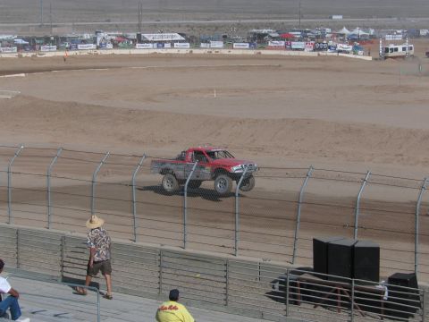 1990 Ford Bronco - red and silver off-road race truck circles the dirt oval as a spectator in a cowboy hat watches from the grandstand rail.