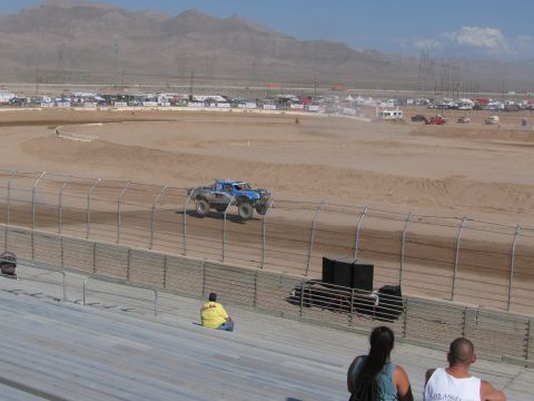 1990 Ford Bronco - blue off-road race truck runs the dirt course while spectators watch from the grandstands under a clear desert sky.