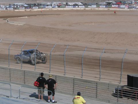1990 Ford Bronco - silver off-road race truck travels the dirt course while two spectators stand watching at the chain-link fence.