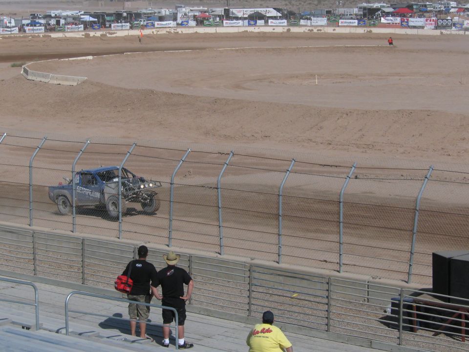 Silver off-road race truck on dirt track with two spectators at the fence