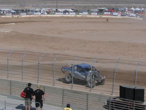 1990 Ford Bronco - blue off-road race truck with exposed rear suspension rounds the dirt track as spectators watch from the fence.