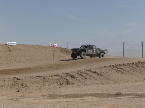 1990 Ford Bronco - black off-road race truck launches off a dirt jump on a desert course lined with sponsor banners and power lines.