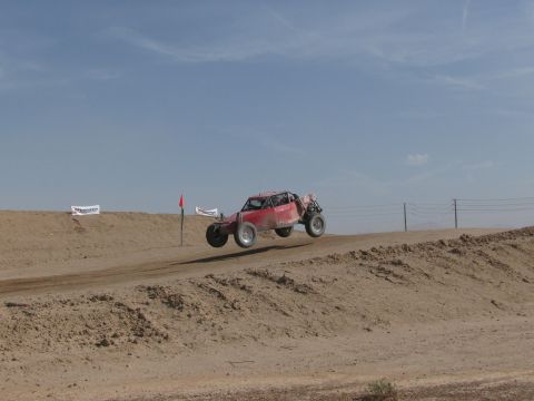 1990 Ford Bronco - red off-road race buggy flies off a dirt jump under a clear blue sky on a desert race course.