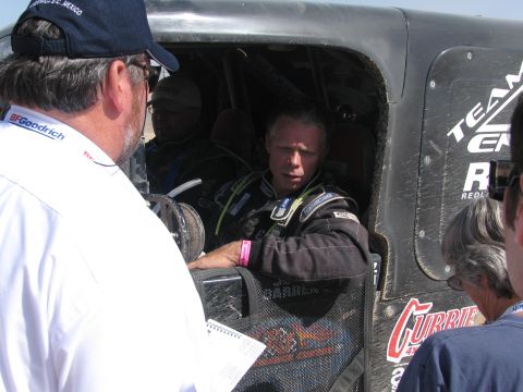 1990 Ford Bronco - driver in a racing fire suit sits in the caged cockpit while an official in a cap talks to him in the pits.