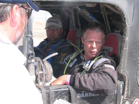 1990 Ford Bronco - Two men in racing suits sit in the roll-caged cockpit of an off-road race vehicle as an official looks on.