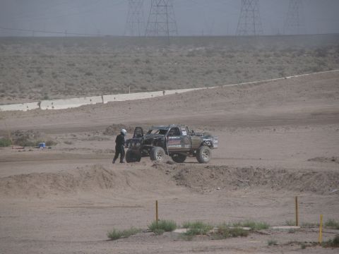 1990 Ford Bronco - off-road race truck sits stopped on a desert course with a helmeted crew member standing beside it near power lines.