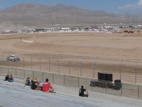 1990 Ford Bronco - off-road race truck rounds the dirt oval track with desert mountains behind as a few spectators watch from the stands.
