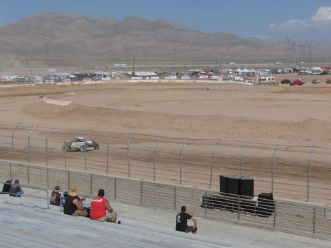 1990 Ford Bronco - light-colored off-road race truck travels the dirt oval below the grandstands with desert mountains on the horizon.