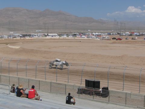 1990 Ford Bronco - silver off-road race truck catches air over the dirt track as spectators watch from the grandstands in the desert.