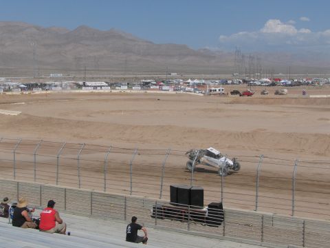 1990 Ford Bronco - silver off-road race truck rounds a dirt oval at a desert short-course track as spectators watch from grandstands beneath distant mountains.