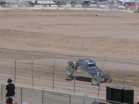 1990 Ford Bronco - blue single-seat off-road race buggy drives along a dirt track seen through chain-link safety fencing at a desert venue.