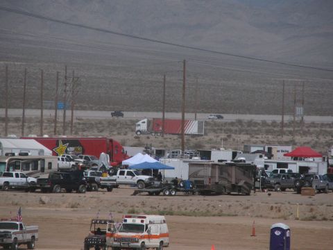 1990 Ford Bronco - crowded race paddock of motorhomes, haulers and an ambulance lines a dusty lot beside a desert highway, with a Rockstar-branded trailer at…