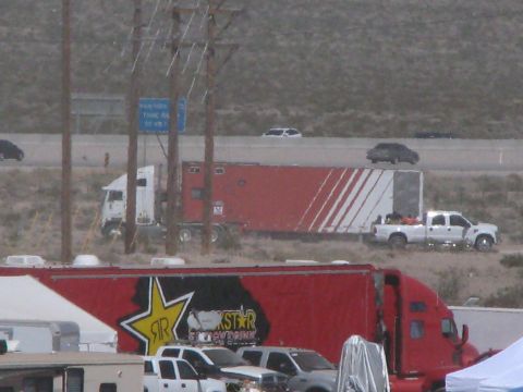 1990 Ford Bronco - Red and white semi-truck race haulers, including a Rockstar Energy Drink trailer, parked beside a highway in a dusty desert lot.