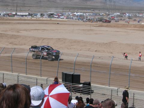 1990 Ford Bronco - dark off-road race truck powers down a dirt straightaway as a packed crowd, one with a red-and-white umbrella, watches from the stands.