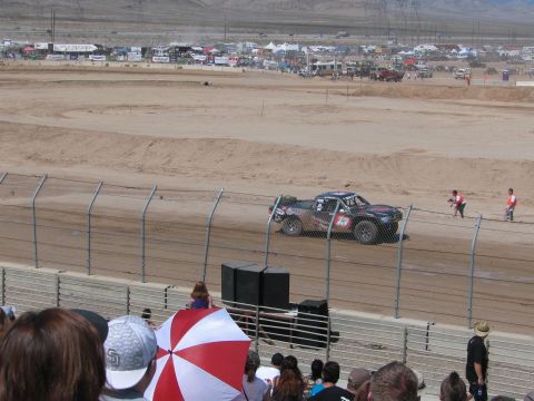 1990 Ford Bronco - off-road race truck travels a dirt track behind safety fencing while spectators and a red-and-white umbrella fill the foreground.